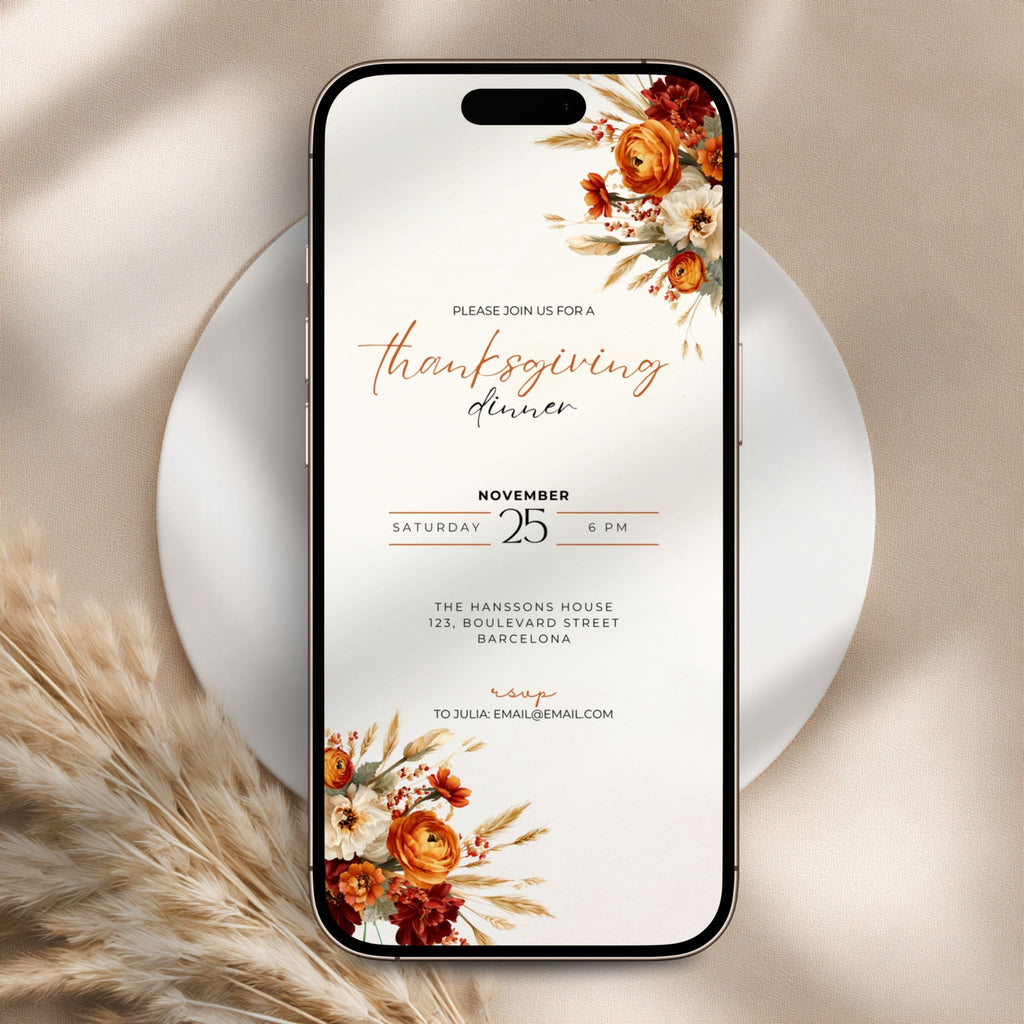 A smartphone centered on a white plate, displaying a Thanksgiving dinner invitation design with floral illustrations in warm autumn tones. The phone is styled on a beige fabric surface with dried grass accents.