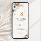 A smartphone case shown from the back on a light neutral background, featuring a Thanksgiving dinner invitation design with floral illustrations, pumpkins, and black text. A dried palm leaf is placed to the left as a styled prop.