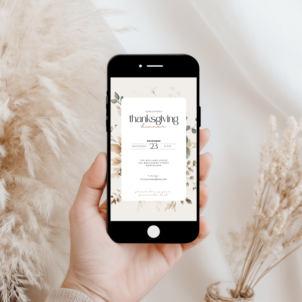 A hand holds a black smartphone displaying a Thanksgiving invitation design with floral accents and elegant typography. The scene is styled with soft beige pampas grass and dried flowers against a bright, clean background.