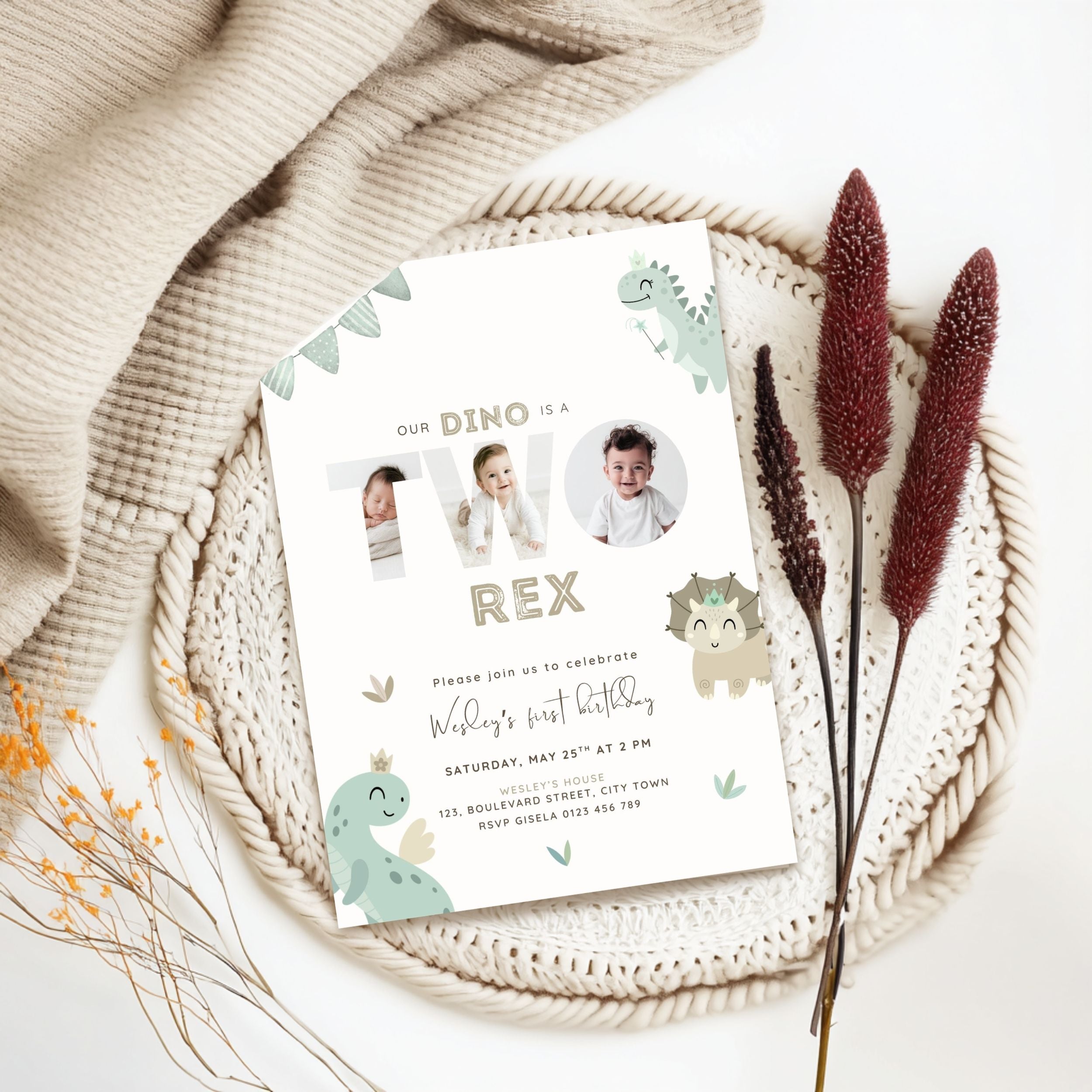 A personalized dinosaur-themed 2nd birthday invitation card styled in a flat lay on a white woven placemat with a beige knit blanket, dried grass stems, and small orange flowers.